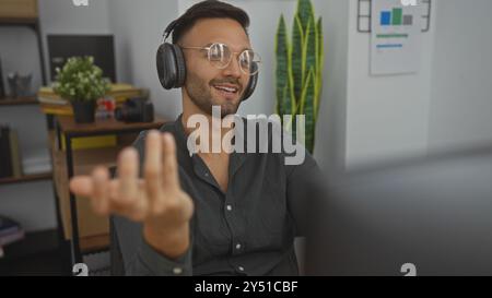 Junger hispanischer Mann mit Bart und Brille, mit Kopfhörern, Gesten, während er in einem Bürozimmer mit Regalen und einer Pflanze saß, in einem Stockfoto