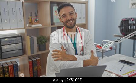 Junger hispanischer Mann mit Bart und überkreuzten Armen lächelnd in einer Tierklinik mit weißem Mantel und Stethoskop, umgeben von medizinischen Büchern und Equ Stockfoto