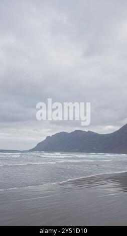 Malerischer Blick auf den Strand von famara mit Wellen, die unter einem bewölkten Himmel auf lanzarote, den kanarischen Inseln, gegen die Sandküste prallen Stockfoto