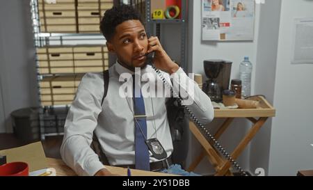 Gutaussehender afroamerikanischer Detektiv, der am Telefon im Polizeirevier spricht, umgeben von Ermittlungsbeweisen. Stockfoto