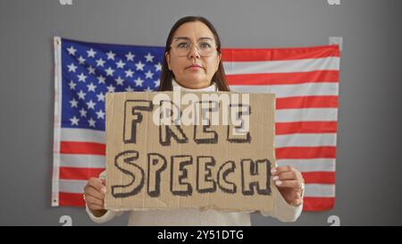 Eine hispanische Frau mittleren Alters, die in einem Innenbereich vor einer amerikanischen Flagge ein Zeichen für die freie Meinungsäußerung hält. Stockfoto