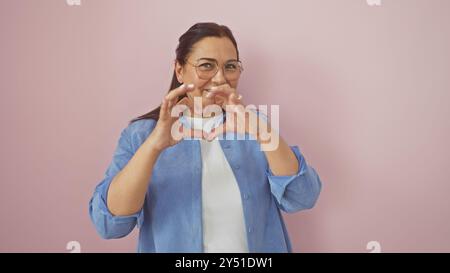 Eine glückliche hispanische Frau mittleren Alters, die eine Herzform vor einem rosa isolierten Hintergrund zeigt Stockfoto