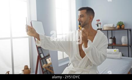 Hübscher junger Mann, der ein Selfie in einem Spa macht, während er ein Hautpflegeprodukt in der Hand hält, einen weißen Bademantel trägt und mit in einem Wellnesscenter drinnen steht Stockfoto