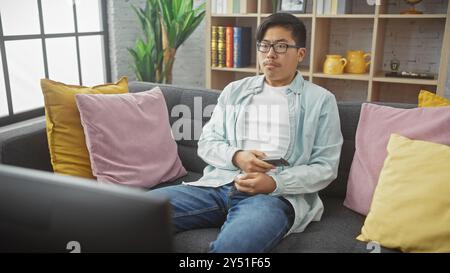 Ein junger asiatischer Mann sitzt lässig auf einer Couch, entfernt in der Hand, in einem gemütlichen Wohnzimmer. Stockfoto