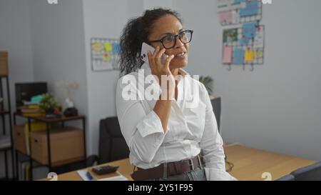 Frau, die auf dem Handy spricht, während sie sich in einem modernen Büro an den Schreibtisch lehnt, mit Anschlagtafel und Bücherregalen im Hintergrund Stockfoto