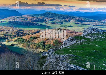 Eine riesige Landschaft mit sanften Hügeln und Herbstwäldern, mit einem felsigen Vorboden und einem entfernten Dorf im Tal. Vitoriano, Araba, Basque Co Stockfoto