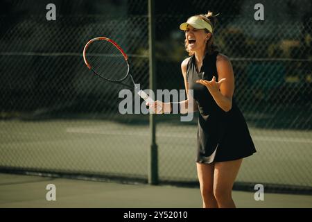 Wütende Frau, die während eines Tennisturniers mit Handgesten und Schläger schreien und dabei Wettkampfgeist und intensive Emotionen zum Ausdruck bringt. Stockfoto