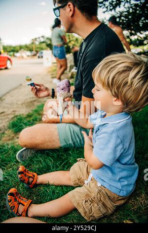 Ein kleiner Junge sitzt auf dem Gras mit einem Erwachsenen, der Eiskegel hält Stockfoto