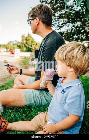 Ein kleiner Junge sitzt auf dem Gras neben einem Erwachsenen und genießt Eis Stockfoto