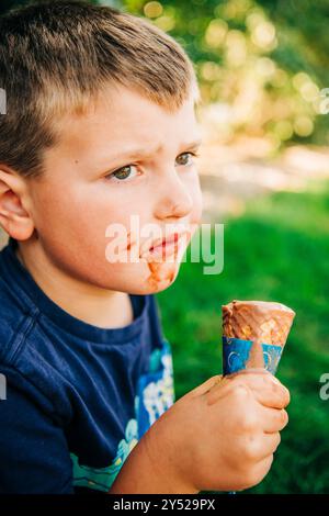 Ein kleiner Junge mit einem schokoladengefärbten Gesicht, der ein schmelzendes Eis hält Stockfoto