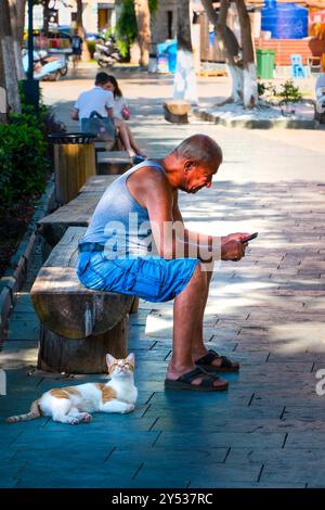 Kas, Antalya, Türkei - 6. August 2024: Ein alter Mann, der bei Sonnenuntergang auf einer Bank sitzt, schaut auf sein Smartphone und eine Katze liegt auf dem Boden neben h Stockfoto