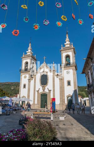 Eine Kirche mit einem großen weißen Turm und einer bunten Blumendekoration, die vom Dach hängt. Die Kirche ist von einem Innenhof mit Bänken und u umgeben Stockfoto