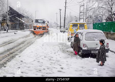 Dieses Bild zeigt eine verschneite Straße in Pahalgam, mit einem Schneepflug, der die Straße räumt, und zwei Kindern, die neben schneebedeckten Autos stehen. Stockfoto