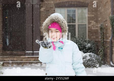 Ein fröhliches junges Mädchen, das warm in Winterkleidung gekleidet ist, genießt den Schneefall vor einem Backsteinhaus. Konzept für Winterferien. Stockfoto