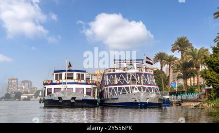 Kairo, Ägypten - 9. Dezember 2023: Nil Kristall Kreuzfahrtschiff legte entlang der Nil Corniche im Bezirk Maadi an. Üppige Palmen und urbane Architektur schaffen eine lebendige Kulisse unter einem klaren blauen Himmel Stockfoto
