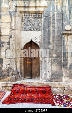 Ein kunstvoller Eingang in einer strukturierten Steinmauer mit komplizierten Schnitzereien, ein lebendiger roter Teppich auf Kopfsteinpflaster unterstreicht den Charme der Altstadt in Baku, Aserbaidschan, während des ersten nationalen Teppichfestes Stockfoto