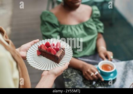 Aus einem hohen Winkel von einer nicht erkennbaren Kellnerin, die einen weißen Teller mit Schokoladenkuchen und Erdbeeren auf der Oberseite hält, während der nicht erkennbare Gast Tee genießt Stockfoto
