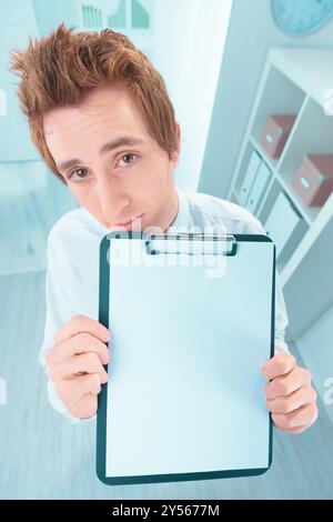Ein fröhlicher Geschäftsmann hält ein leeres Klemmbrett über dem Kopf und macht ein lustiges Gesicht in einem Firmenbüro. Passen Sie den Bereich mit Text für eine PL an Stockfoto
