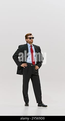 Ein harfengekleideter Mann passt seine Sonnenbrille an, trägt rote Krawatte und eine zweireihige Jacke und sieht vor weißem Studiohintergrund selbstbewusst aus. Stockfoto