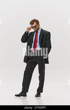 Harfengekleideter Mann, der seine Sonnenbrille anpasst, rote Krawatte und zweireihige Jacke trägt und selbstbewusst vor weißem Studiohintergrund blickt. Stockfoto