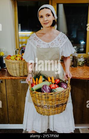 Die Frau trägt ein traditionelles weißes Kleid und Schürze und hält einen großen Korb mit frischem Gemüse, einschließlich Karotten, Tomaten und Eierlikör Stockfoto