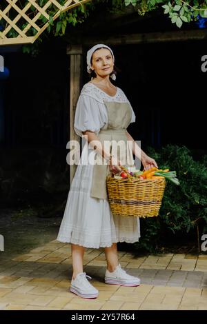 Die Frau trägt ein traditionelles weißes Kleid und Schürze und hält einen großen Korb mit frischem Gemüse, einschließlich Karotten, Tomaten und Eierlikör Stockfoto