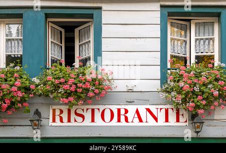 Italienisches Restaurantschild an der Holzfassade eines Hauses mit Fenstern und blühenden Blumen Stockfoto