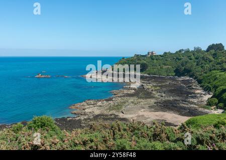 Ein malerischer Blick auf eine felsige Küste in der Bucht von Le Sauchet mit einem Gebäude auf einem grünen Hügel mit Blick auf das türkisfarbene Meer an klaren Tagen. Stockfoto