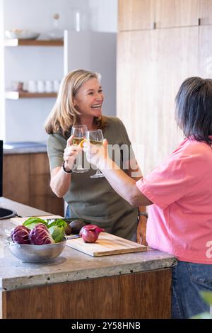 Zu Hause, mit Weingläsern zu toasten, zwei ältere, multirassische Freundinnen zusammen in der Küche Stockfoto