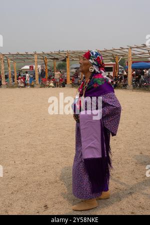 Die weibliche Powwow-Tänzerin mittleren Alters trägt ein lila Kleid und farbenfrohe Tücher, während sie einen lila Schal trägt. Fotografiert in MT Stockfoto