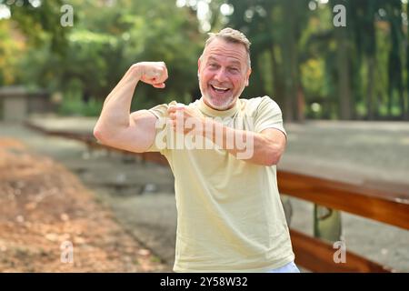 Ein glücklicher reifer Mann, der seinen Arm beugt und mit einem großen Lächeln auf seinen Bizeps zeigt, während er einen unterhaltsamen und aktiven Moment im Freien in einem sonnigen Park genießt. Stockfoto