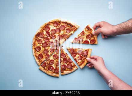 Mann und Frau greifen sich Pizzascheiben. Geschnittene Peperoni-Pizza auf blauem Hintergrund, Ansicht oben. Blick von oben mit köstlicher Peperoni-Pizza Stockfoto