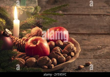 Weihnachtslandschaft mit einer Holzplatte voller Walnüsse, Haselnüsse und Äpfel, grünen Tannenzweigen und einer Geschenkbox, auf einem Vintage-Tisch bei Kerzenschein Stockfoto