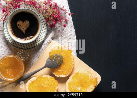 Frühstück mit einer Tasse Kaffee mit Schaumherz, mit Pfirsichmarmelade bestrichenen Brotscheiben auf einem Schneidebrett, umgeben von Blumen, auf einem schwarzen Tisch Stockfoto