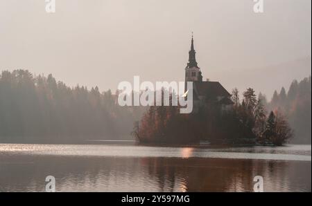 Landschaft bei Sonnenaufgang mit der Kirche der Gottesmutter am Bleder See in Slowenien. Neblige Landschaft mit dem Bleder See an einem sonnigen Tag im Dezember Stockfoto