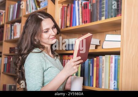 Junge schöne Frau, lässig gekleidet, zufrieden mit dem Buch, das sie aus der Bibliothek nahm, während sie auch eine Tasse Kaffee oder Tee in der Hand hielt Stockfoto