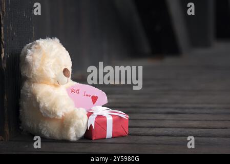 Geschenkfoto mit einem niedlichen Teddybär, der eine Papiernote mit den Worten With Love hält, und einer roten Geschenkbox in einem Vintage-Holzdekor Stockfoto