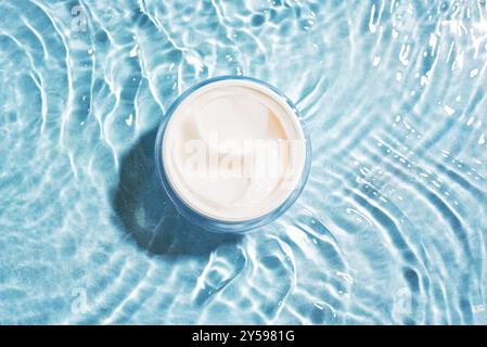 Geöffnetes Glas Kosmetikcreme auf klarem, blauem Wasser, Draufsicht. Feuchtigkeitsspendendes Kollagen-Schönheitsprodukt, beruhigende Gesichts- oder Körpercreme. Stockfoto