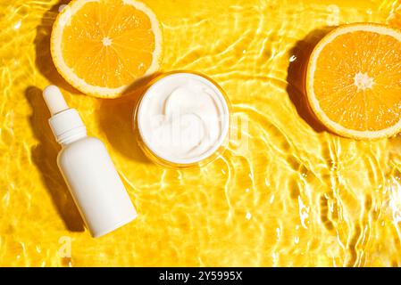 Weiße Kosmetikcreme und Serumflasche ohne Marke auf gelbem Wasserhintergrund mit orangen Zitrusscheiben. Feuchtigkeitsspendendes Vitamin C Schönheitsprodukt, Bio s Stockfoto