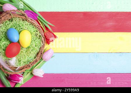 Oster-Banner-Konzept mit einem Korb gefüllt mit dekorativem Gras und bemalten Eiern, umgeben von Tulpen, auf einem mehrfarbigen Holzhintergrund Stockfoto