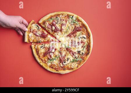 Flacher Lay mit der Hand einer jungen Frau, die sich ein Stück Pizza schnappt. Hausgemachte Pizza mit Schinken Crudo und Rucola. Leckere Schinken Pizza Stockfoto