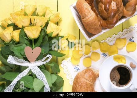 Morgenmahlzeit mit Gebäck, einer Tasse aromatischem Kaffee und einem großen Strauß von gelben Rosen und Blütenblättern auf einem gelben Holztisch Stockfoto