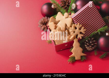 Geschenkbox mit Honig- und Ingwerkeksen, roten Kugeln, Tannenzapfen und Tannenzweigen auf rotem Hintergrund. Weihnachtsbanner, Weihnachtsbacken und Geschenke Stockfoto