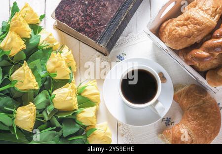 Frühstückstisch mit einer Tasse Kaffee, frisch gebackenen Croissants, einem alten geschlossenen Buch und einem großen Strauß gelber Rosen auf einem weißen Holztisch Stockfoto