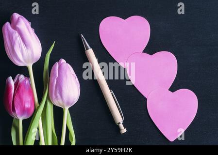 Farbenfrohe Tulpen, ein Holzstift und drei rosafarbene herzförmige Papierpfosten mit Platz für Nachrichten auf schwarzem Holzhintergrund Stockfoto