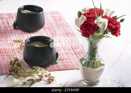 Strauß aus weißen Rosen und roten Nelken in einer niedlichen Vase und zwei schwarzen Tassen Tee mit getrockneten Lindenblüten, auf einem weißen Tisch, im Sonnenlicht Stockfoto