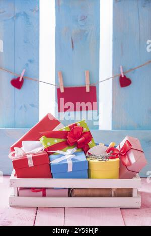 Bunte Geschenkboxen in einer weißen Holzkiste auf einem rosa Tisch mit roter Papierkarte und Herzen, die an eine Schnur gebunden sind, auf einem blauen Zaun Stockfoto