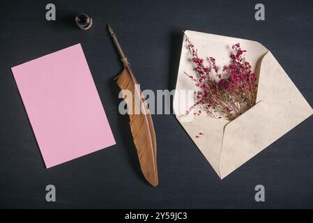 Vintage-Umschlag voll mit rosa Blumen, einem antiken Federstift, einem Tintenbehälter und einer leeren Nachrichtenkarte auf schwarzem Holzhintergrund Stockfoto