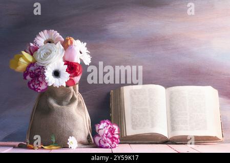 Wunderschöner Strauß aus Rosen, Gerbera, Tulpen und Nelken auf einem rosafarbenen Tisch und ein antikes offenes Buch, das vor einem lila hölzernen Hintergrund gelehnt wurde Stockfoto