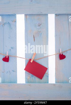 Ungeschriebene rote Papierkarte, umgeben von roten Herzen, gebunden an eine Leinenkette mit Holzklammern, auf einem blauen Zaun Stockfoto